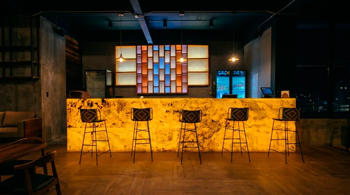 Vista frontal de una barra de bar de diseño con revestimiento retroiluminado StoneFlex Indian Autumn en tono dorado ámbar y vetas oscuras, integrando texturas naturales y mobiliario moderno.